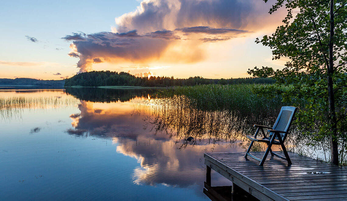 Valoisa kesäilta keskiyöllä järven rannalla Etelä-Suomessa, kuuropilvi heijastuu tyyneen järveen ja laiturilla on yksi aurinkotuoli